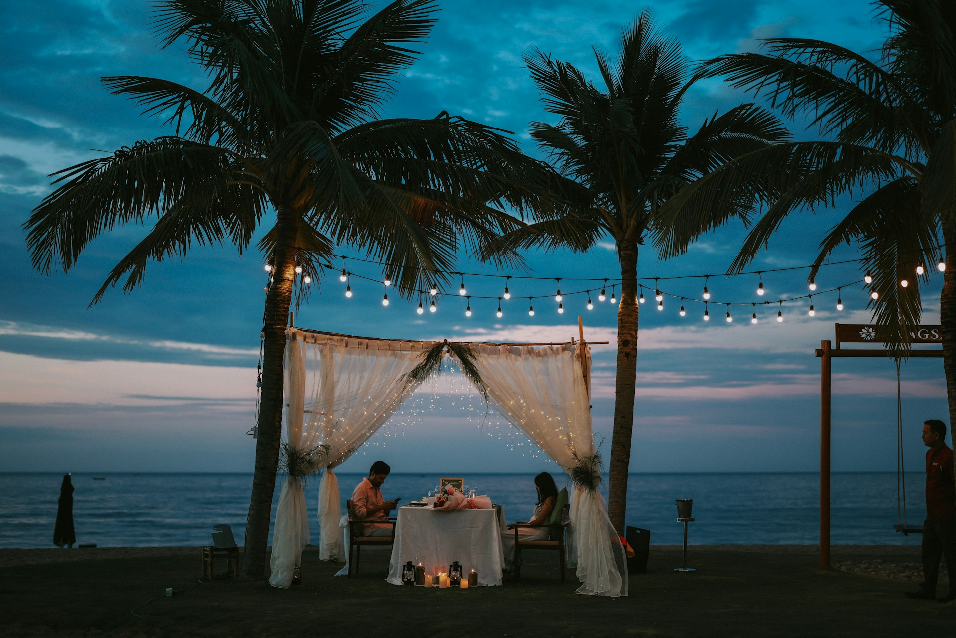 Couple dining under palm trees at dusk with string lights.